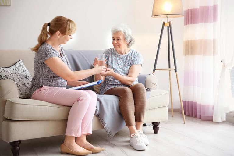 Caregiver in a grey top handing a glass of water to a smiling older woman on a sofa at home
